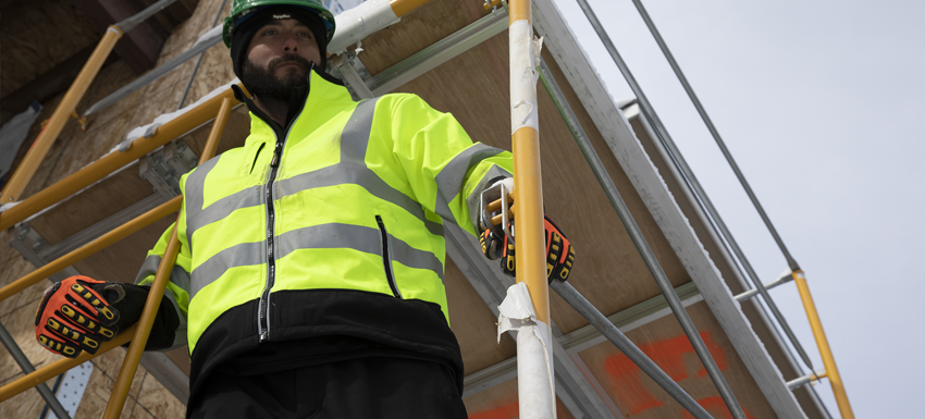 A man working in construction wearing a HiVis lime jacket, HiVis orange gloves and a safety helmet stands on scaffolding and looks into the distance.