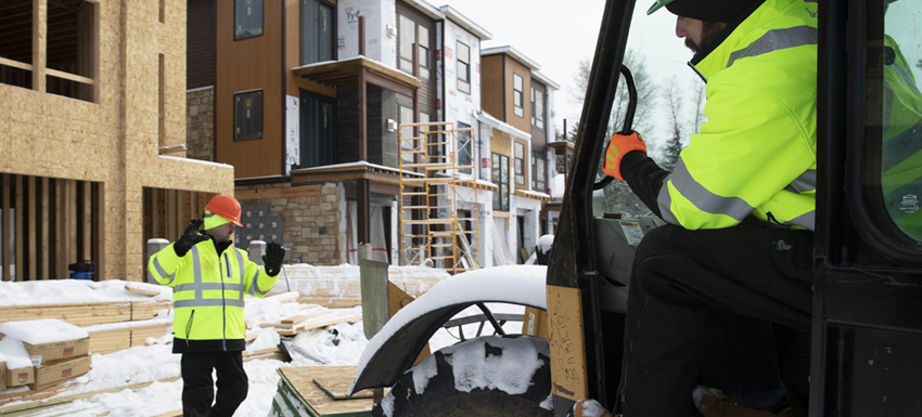 Two construction workers in HiVis lime jackets and orange helmets working on the construction of a house. One man directs the other, who’s driving a forklift.